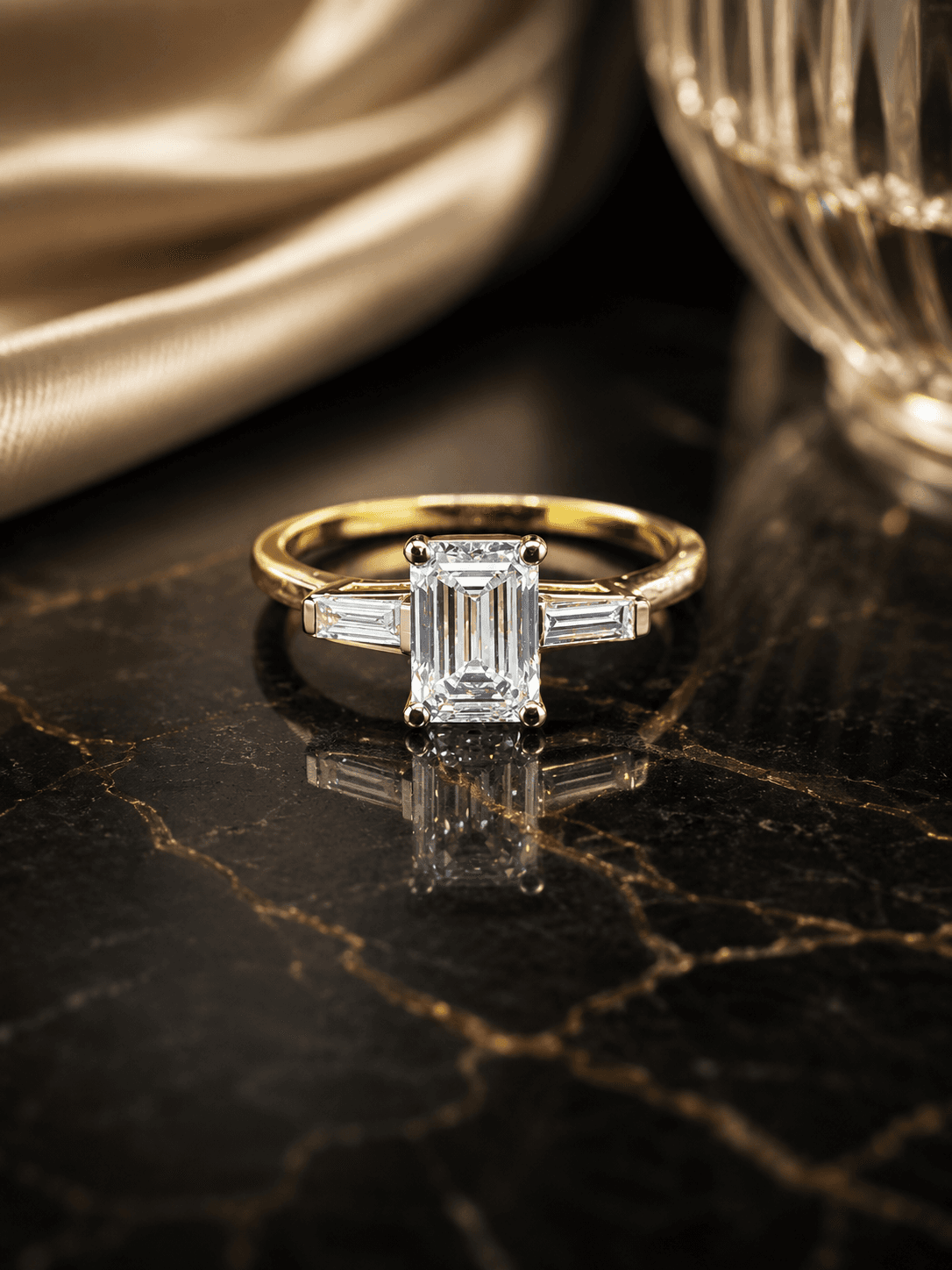 Emerald-cut lab-grown diamond three-stone ring with tapered baguette side stones in yellow gold, on dark marble.