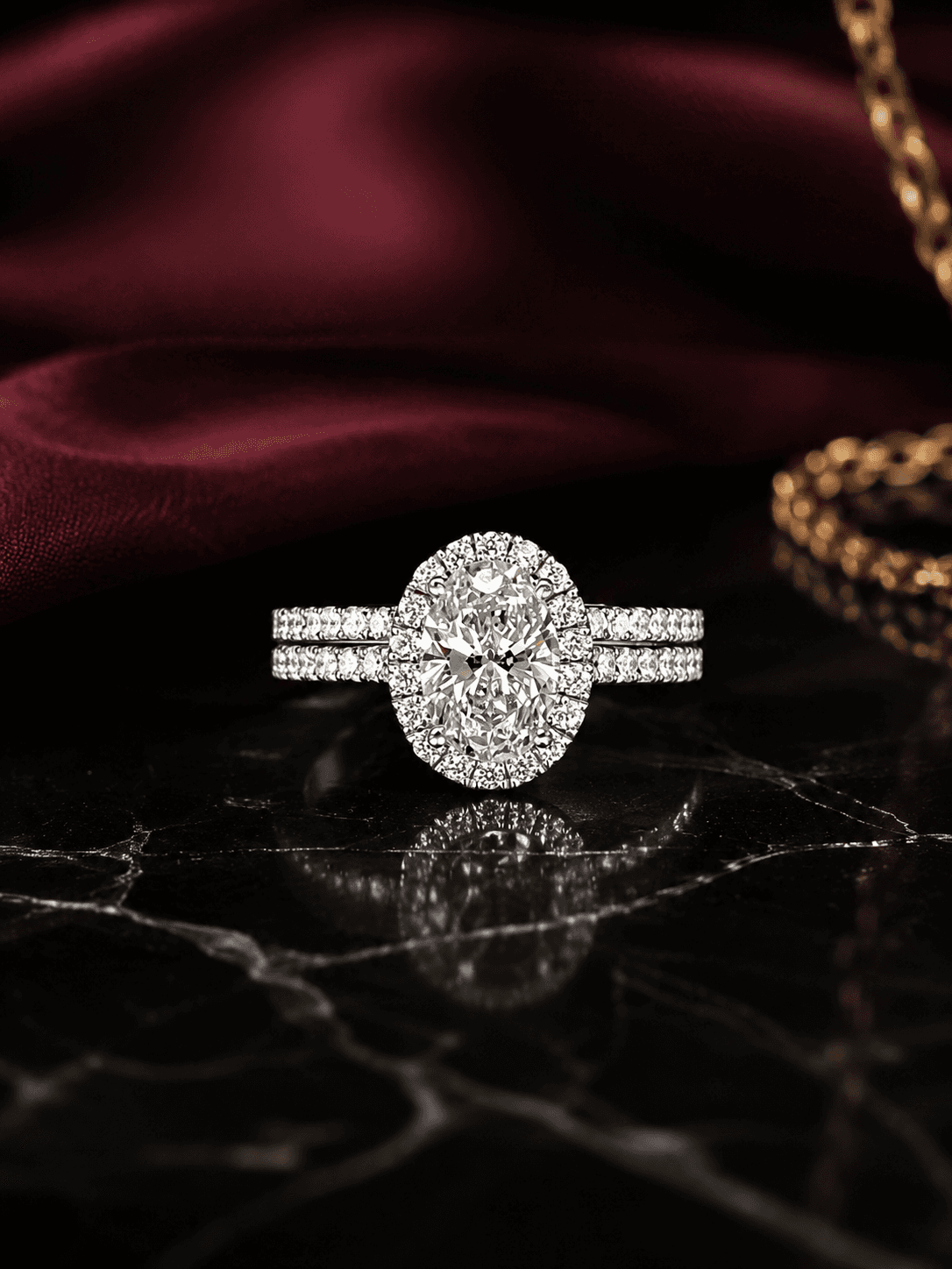 Oval lab-grown diamond halo engagement ring with a matching pavé wedding band in white gold, on black marble with burgundy silk.