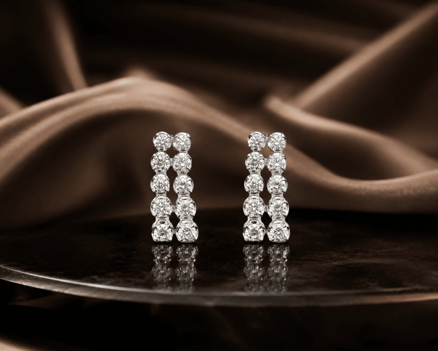 A pair of double-row round lab-grown diamond huggie earrings in white gold, on a dark tray against brown silk.